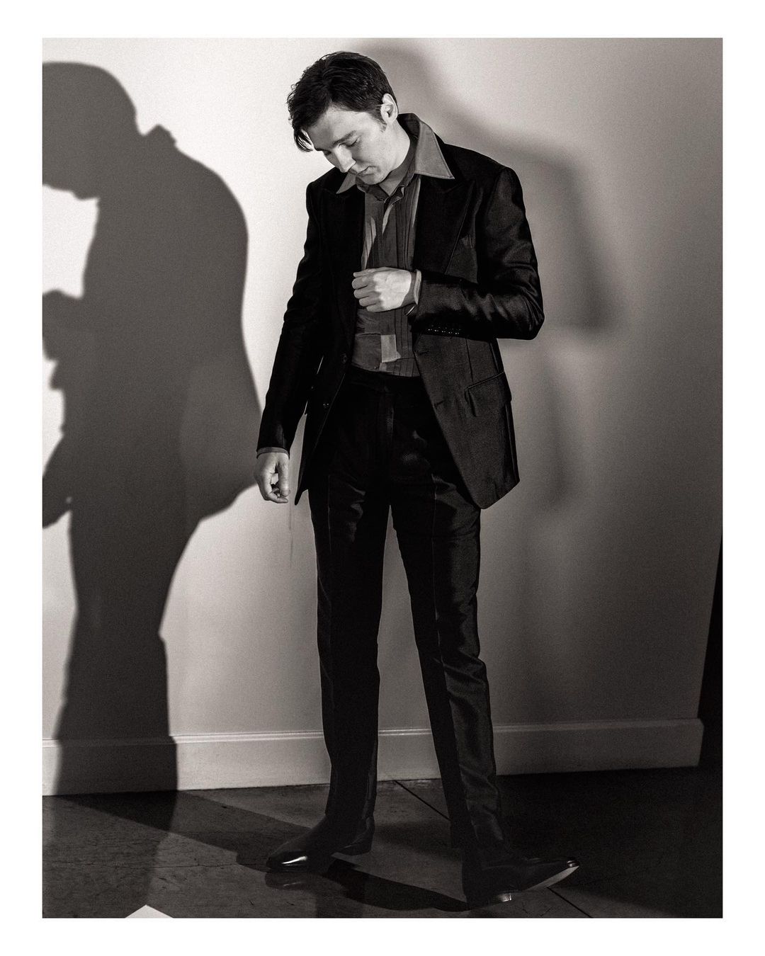 Monochrome full-length portrait of a man in a suit standing indoors against a wall with a shadow, wearing a button-down shirt.