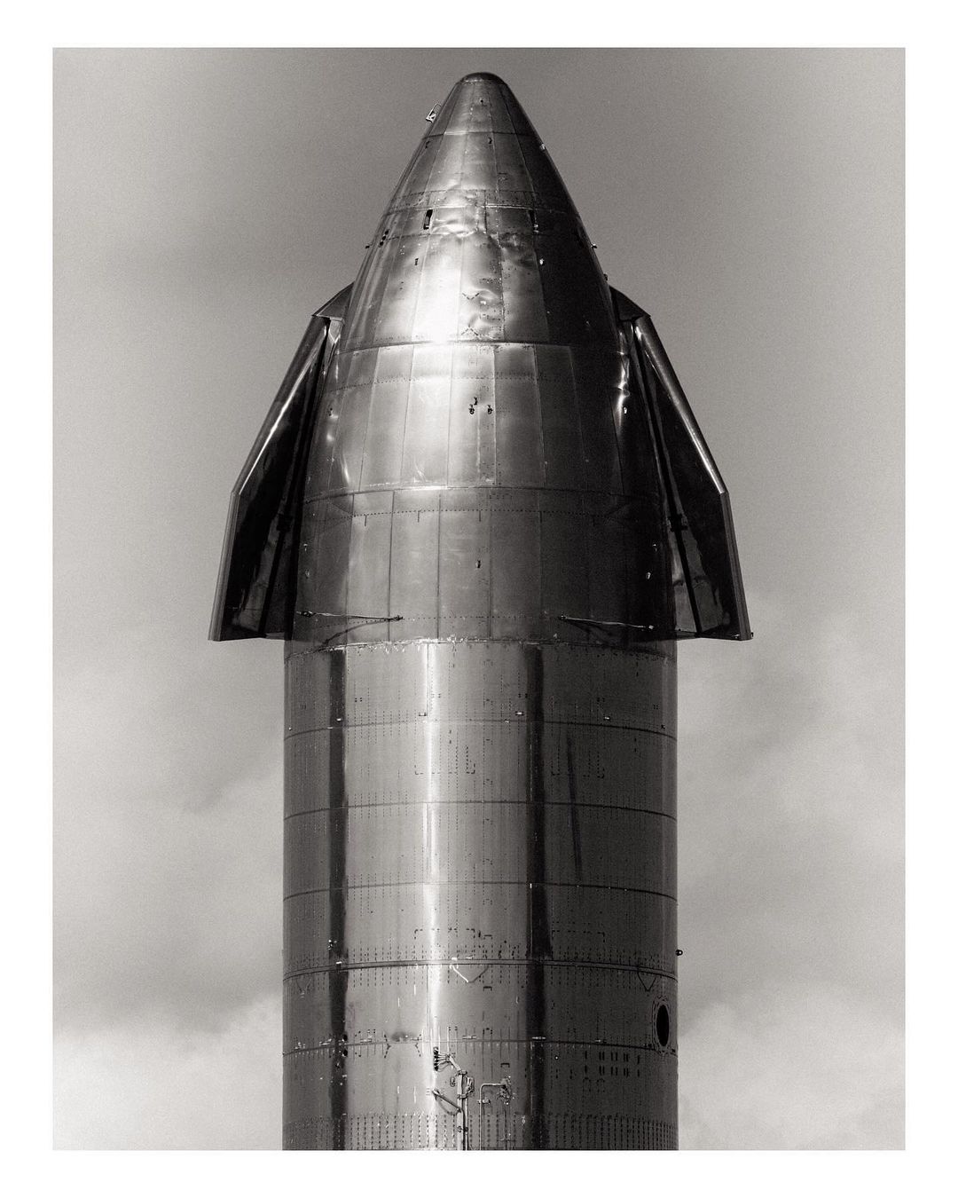 Monochrome photograph of a metallic space rocket. The rocket tapers to a point at the top with wing-like structures at mid-point.