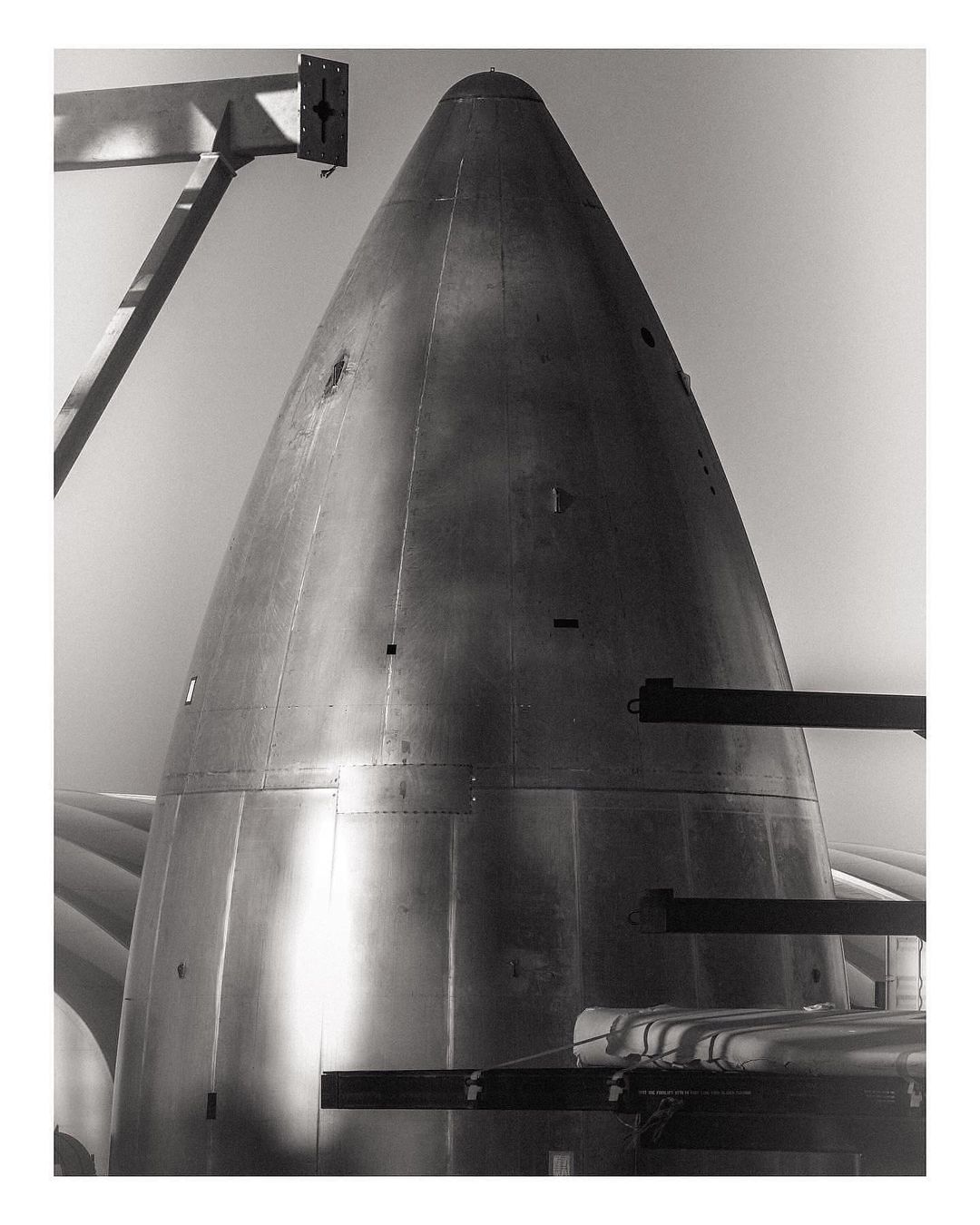 Monochrome photograph of a rocket or missile nose cone under construction, with support beams against a sky background.