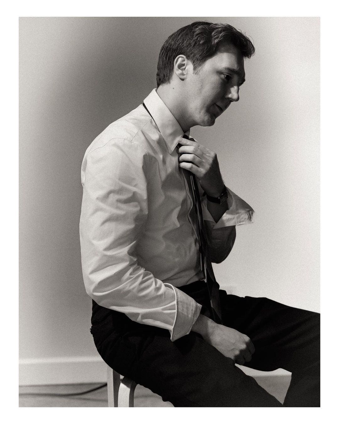 Black and white portrait of a man in a dress shirt and tie sitting indoors, adjusting his tie, captured with natural lighting.