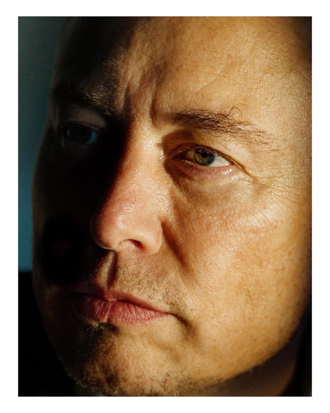 Close-up portrait of an adult male with short hair. The image features dramatic lighting and shadows across the face, highlighting skin texture.