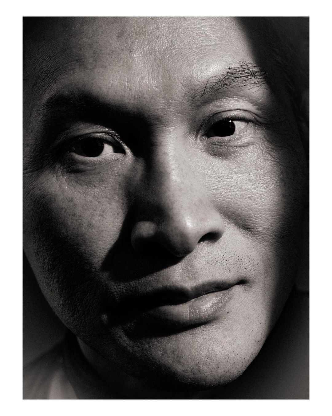 Monochrome close-up portrait of an Asian adult male with a partial face shadow, smiling slightly, and facing the camera in a photo studio.