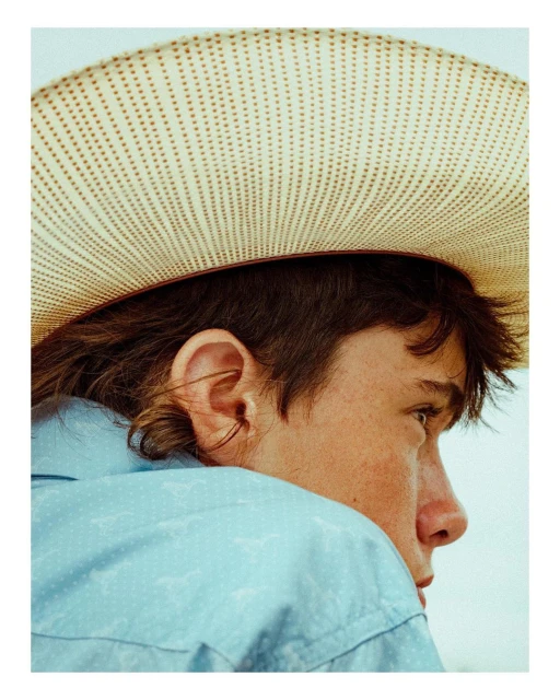 Profile shot of a young adult male with a mullet haircut wearing a cowboy hat and light blue patterned shirt in a brightly lit setting.