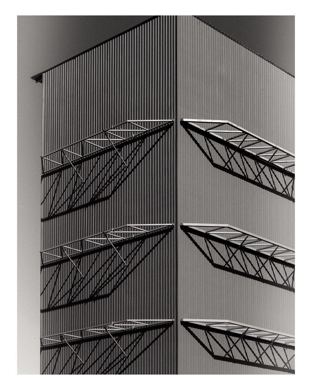 Architectural black and white photograph of a building corner with steel siding and window awnings casting geometric shadows.