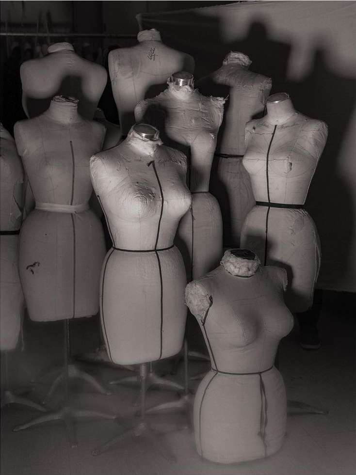 Monochrome photo of a group of dress forms on stands, arranged closely together in a studio setting. Soft lighting. 