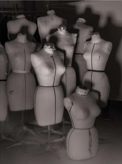 Monochrome photo of a group of dress forms on stands, arranged closely together in a studio setting. Soft lighting. 