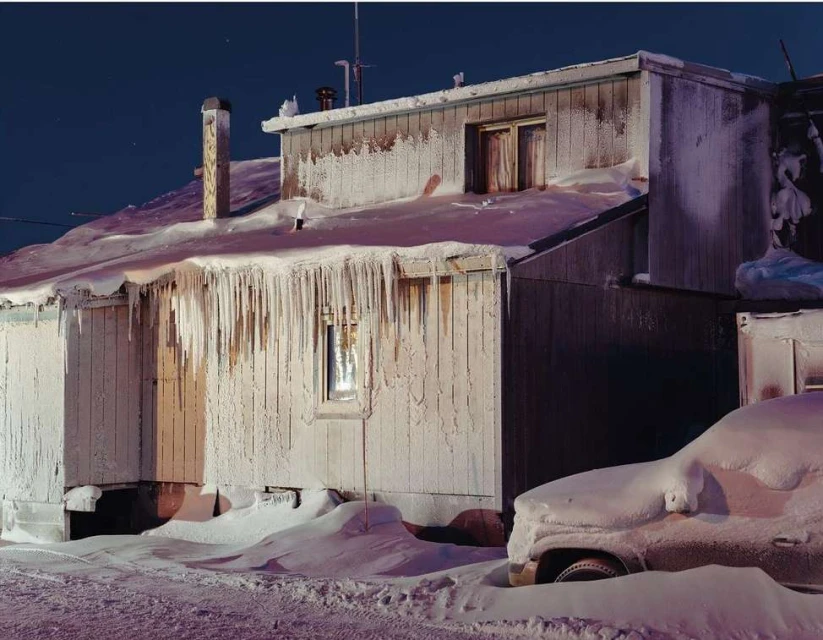 Snow-covered house with large icicles hanging from the roof, illuminated by streetlights under a dark blue evening sky, with a snow-covered car.