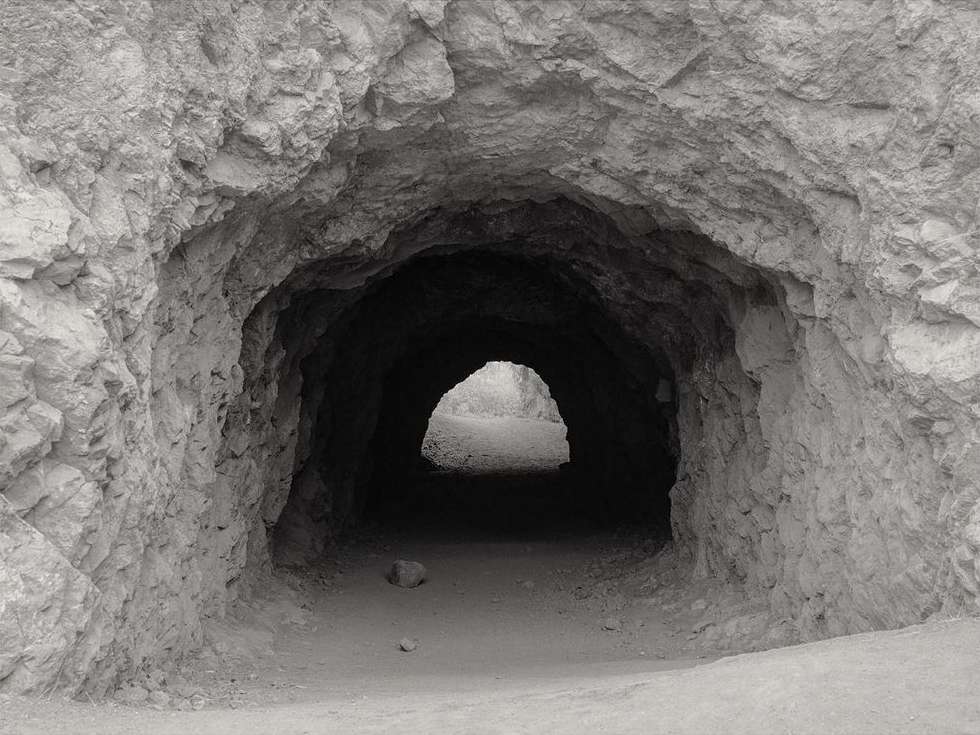 Monochrome shot of a dark tunnel entrance with rough rock walls, leading to a brighter opening in the distance.