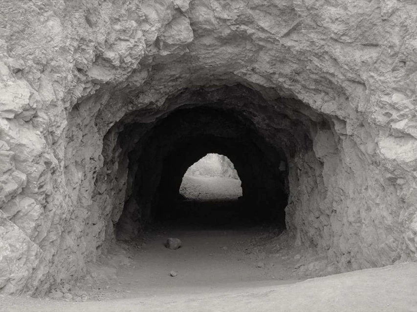 Monochrome shot of a dark tunnel entrance with rough rock walls, leading to a brighter opening in the distance.