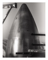 Monochrome vertical photograph of a large metallic rocket nose cone with structural scaffolding elements surrounding it.