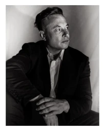 Monochrome portrait of Elon Musk wearing a dark suit jacket and white shirt, looking away from the camera.