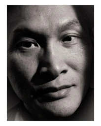 Close-up black and white portrait of an Asian man with textured skin, sharp lighting, and a neutral expression.