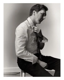 Portrait of Paul Dano adjusting his tie while seated in a white shirt and dark trousers against a neutral background.