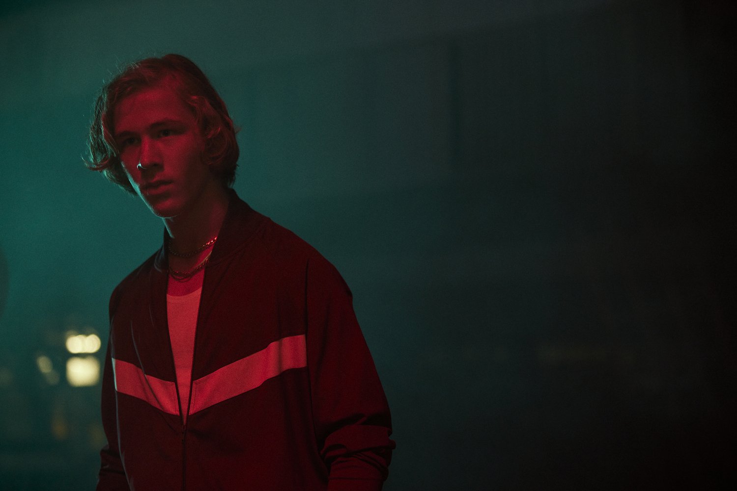 Portrait of a young adult man with blond hair in a black jacket with red stripes, posing against a dark background with red and green lighting.