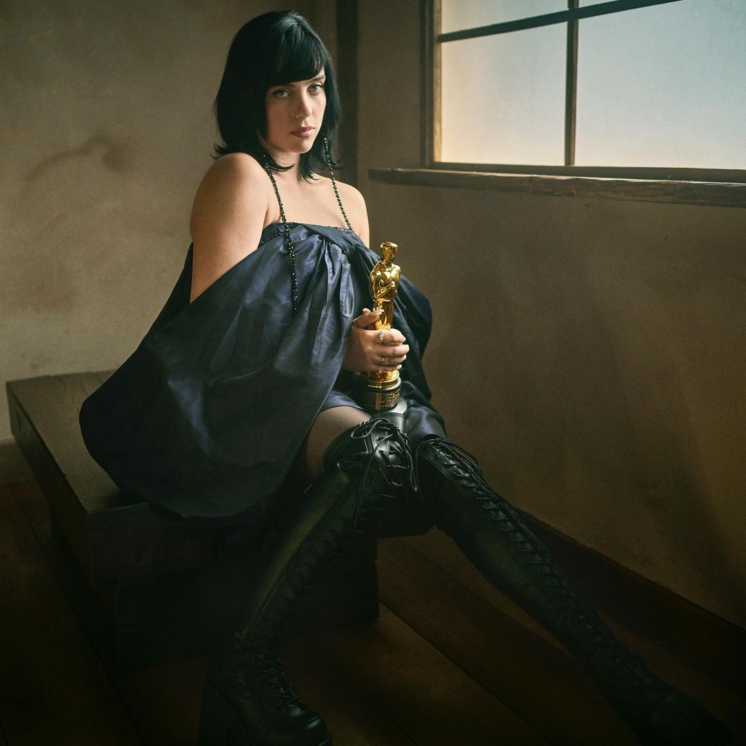 Brunette woman in black dress and boots holds an Oscar statuette indoors, near a window with natural light, sitting on a wooden surface.