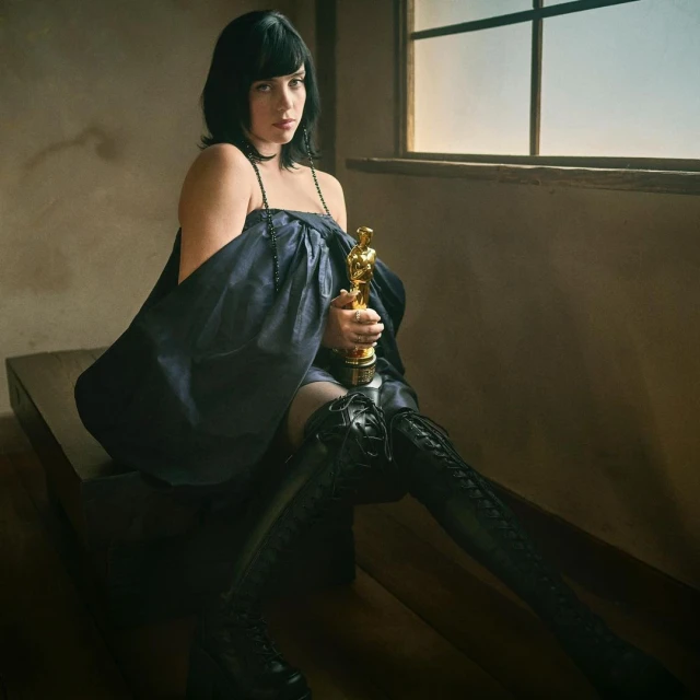 Brunette woman in black dress and boots holds an Oscar statuette indoors, near a window with natural light, sitting on a wooden surface.