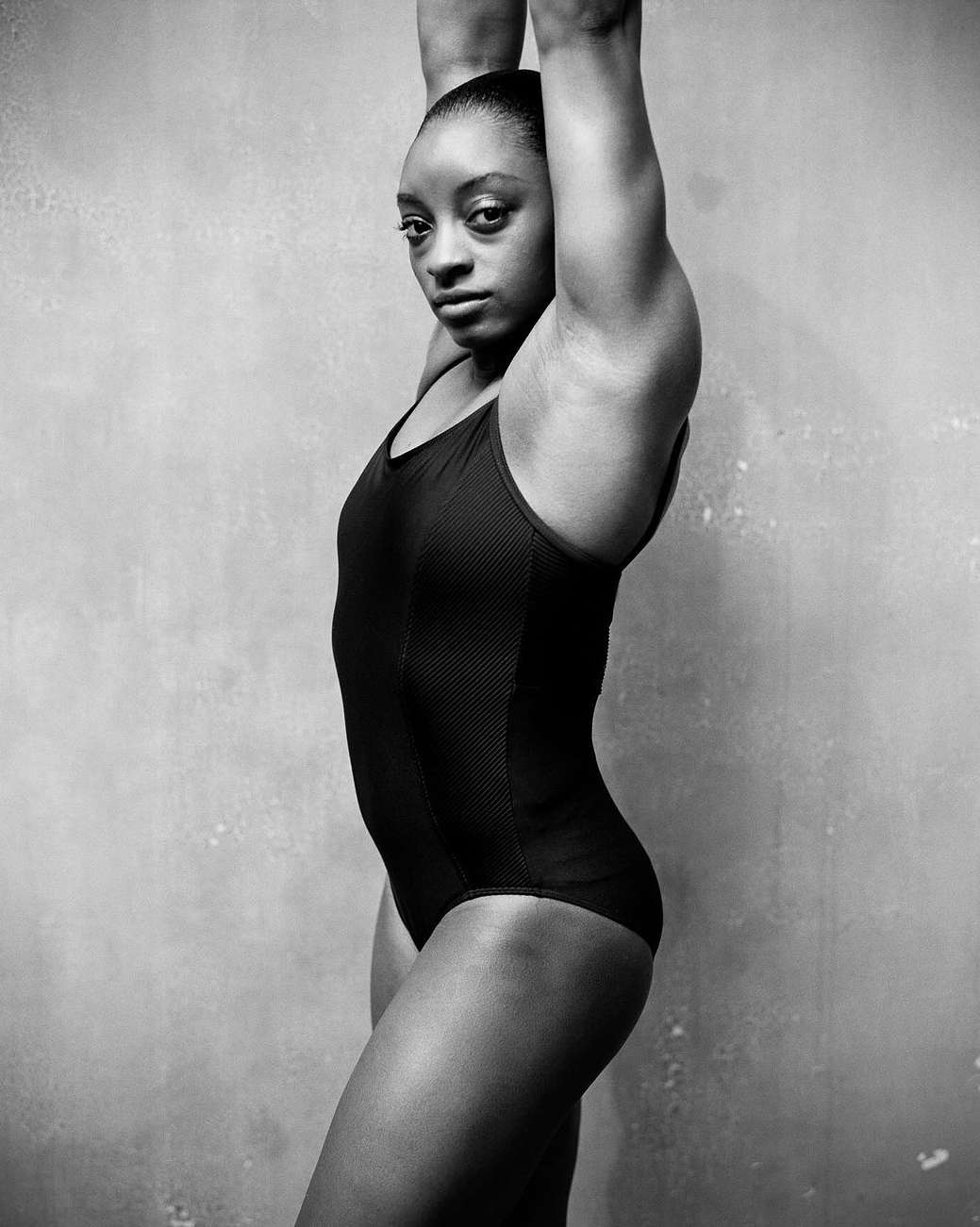 Profile portrait of an athletic African-American adult female in a black leotard, arms raised, set against a textured gray backdrop.