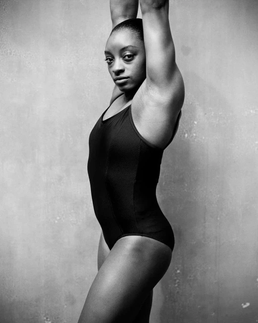 Profile portrait of an athletic African-American adult female in a black leotard, arms raised, set against a textured gray backdrop.