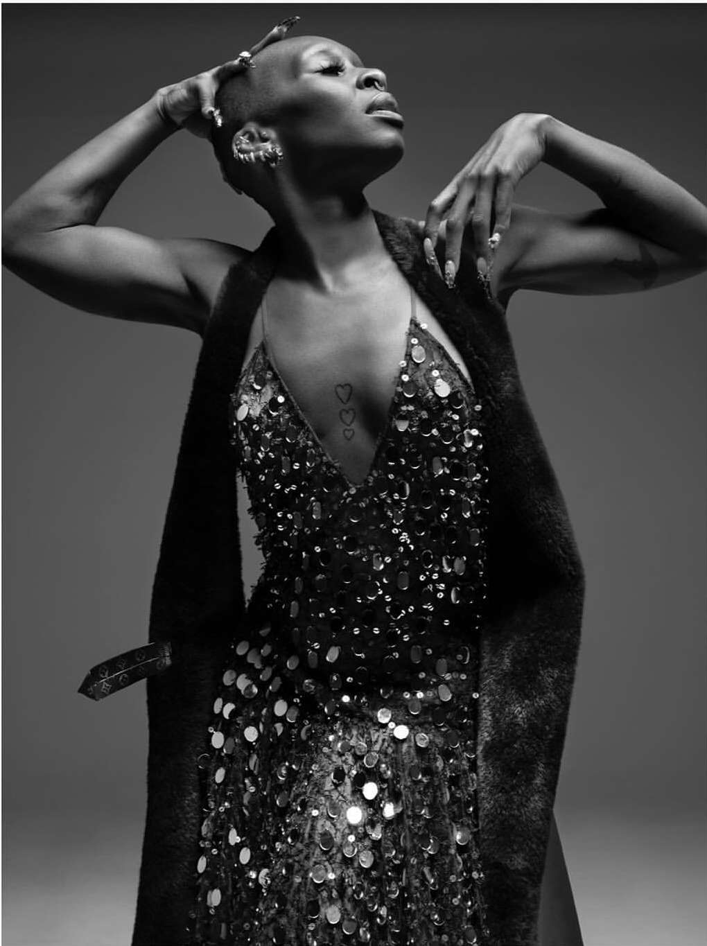 Black and white portrait of an adult woman with short hair wearing a sequined dress and a fur vest, looking up with hands raised.