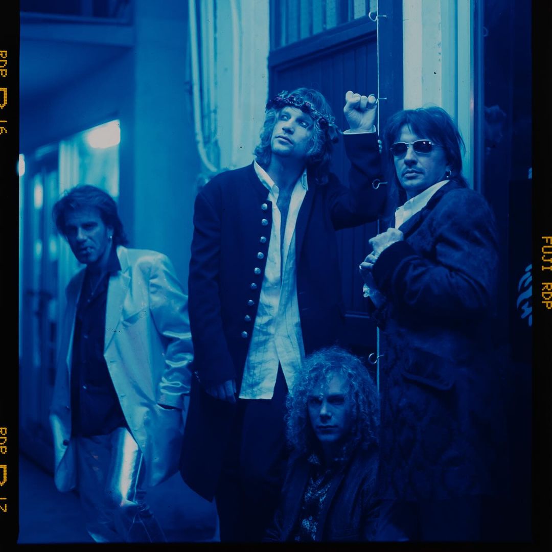 Indoor shot of four adult men in dark jackets, one with long curly hair, looking at the camera in a dramatic blue color grade.