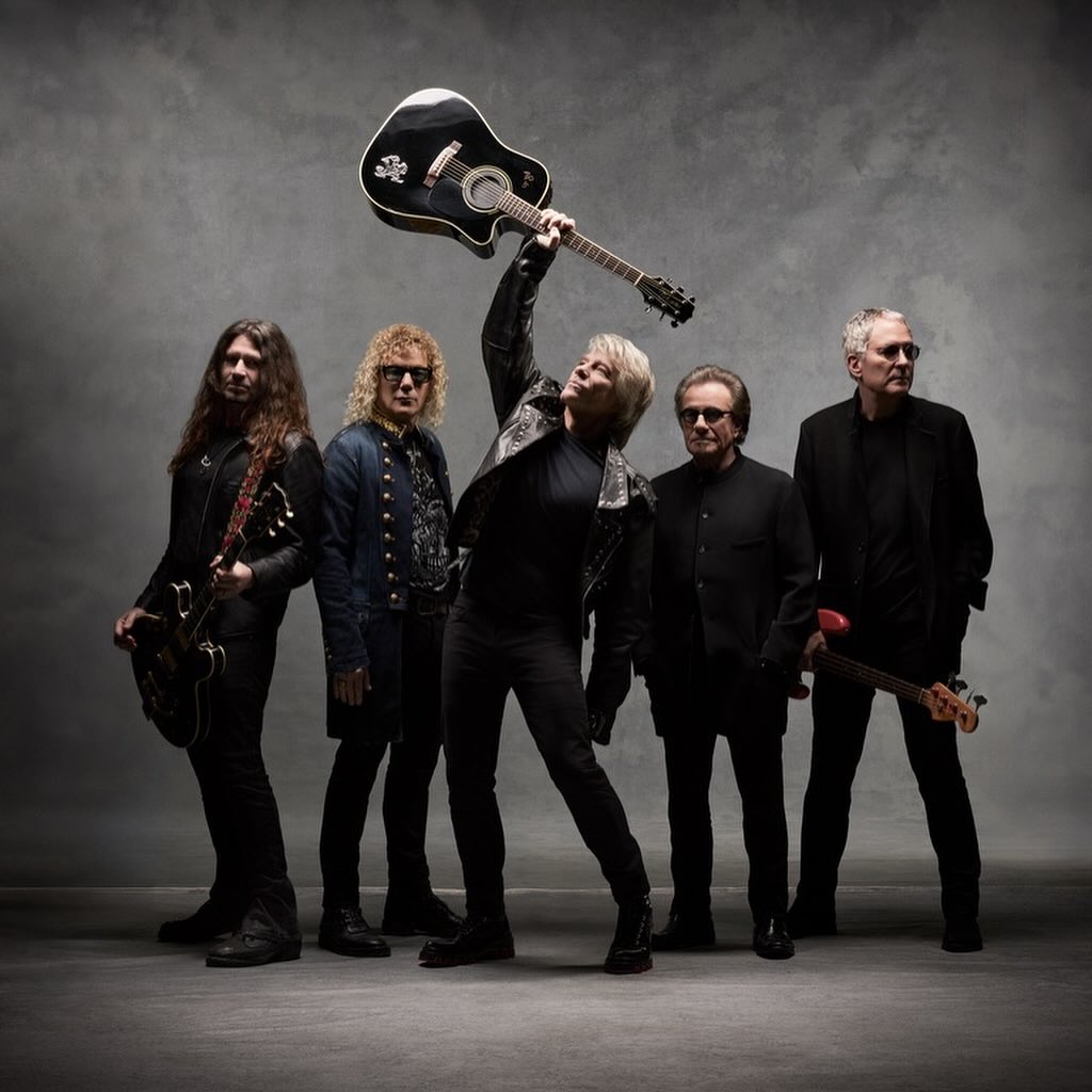 Band poses together in photo studio with instruments and all-black outfits. One man raises his guitar above his head.