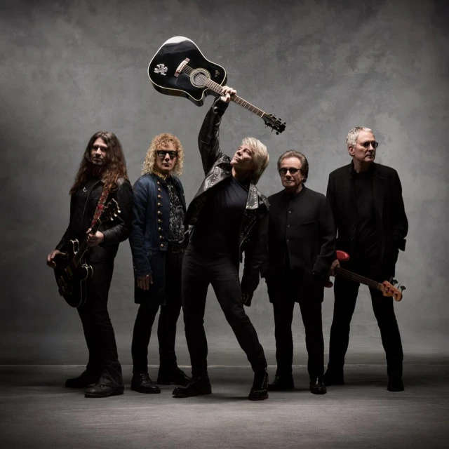 Band poses together in photo studio with instruments and all-black outfits. One man raises his guitar above his head.