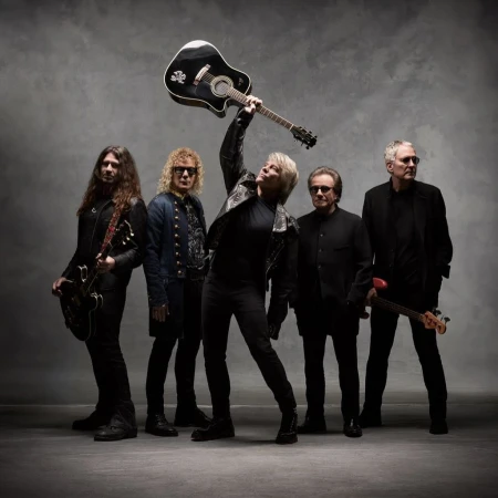 Band poses together in photo studio with instruments and all-black outfits. One man raises his guitar above his head.