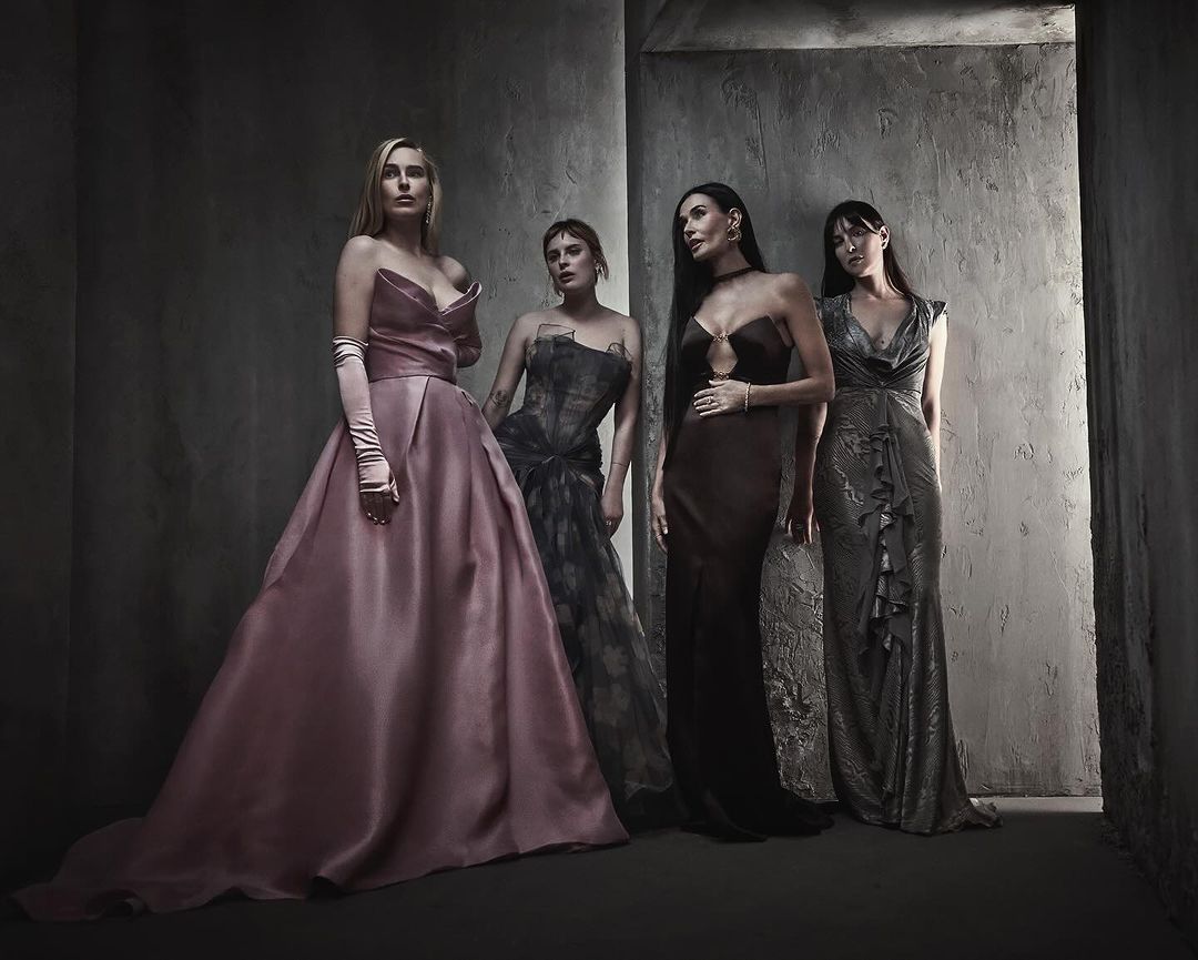 Four women in formal gowns stand together in a dramatic indoor setting with gray textured walls, posing for a high-fashion studio portrait.