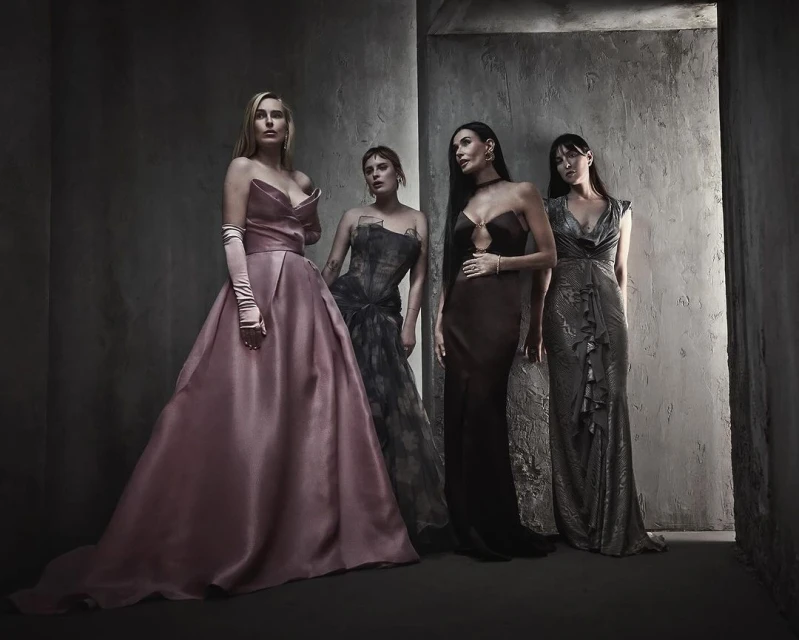 Four women in formal gowns stand together in a dramatic indoor setting with gray textured walls, posing for a high-fashion studio portrait.