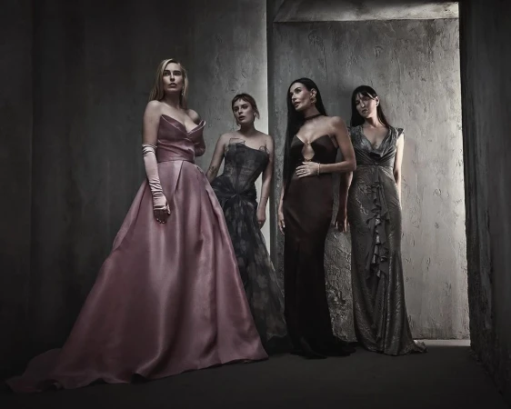 Four women in formal gowns stand together in a dramatic indoor setting with gray textured walls, posing for a high-fashion studio portrait.