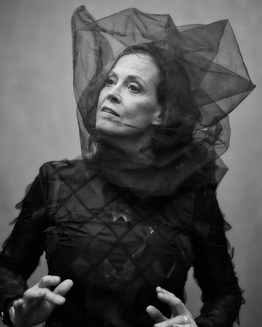 Monochrome portrait of an adult woman with a sheer black head covering, wearing a dark textured top, against a solid gray background.