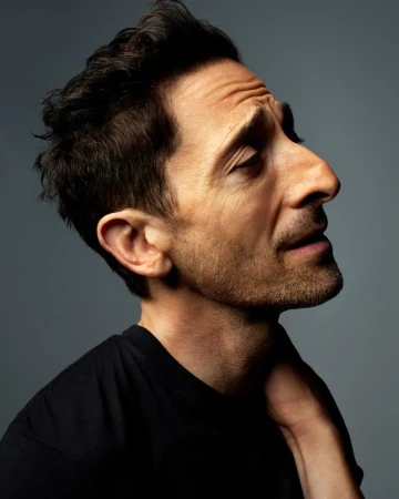 Profile portrait of a man with dark hair wearing a black t-shirt against a gray background. He is looking up with a slight grin.