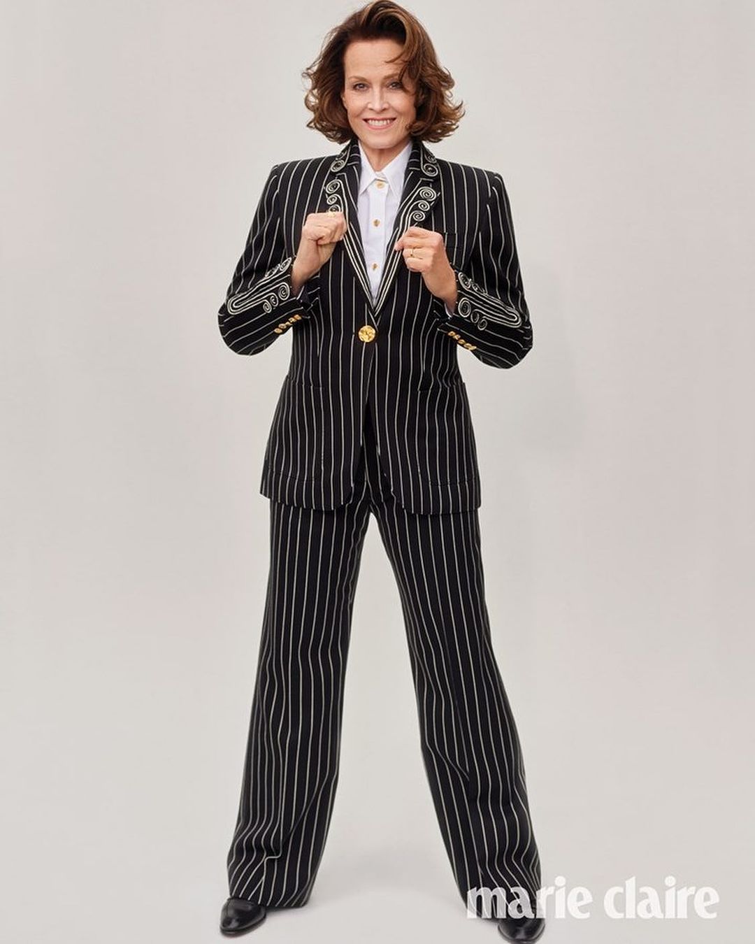 Full shot of Sigourney Weaver in a striped suit and white shirt, standing in a studio with a neutral background, smiling, and holding her hands up.