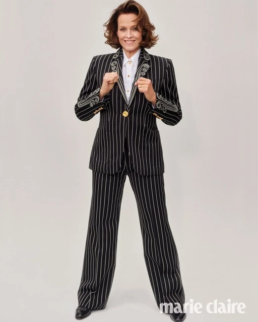 Full shot of Sigourney Weaver in a striped suit and white shirt, standing in a studio with a neutral background, smiling, and holding her hands up.