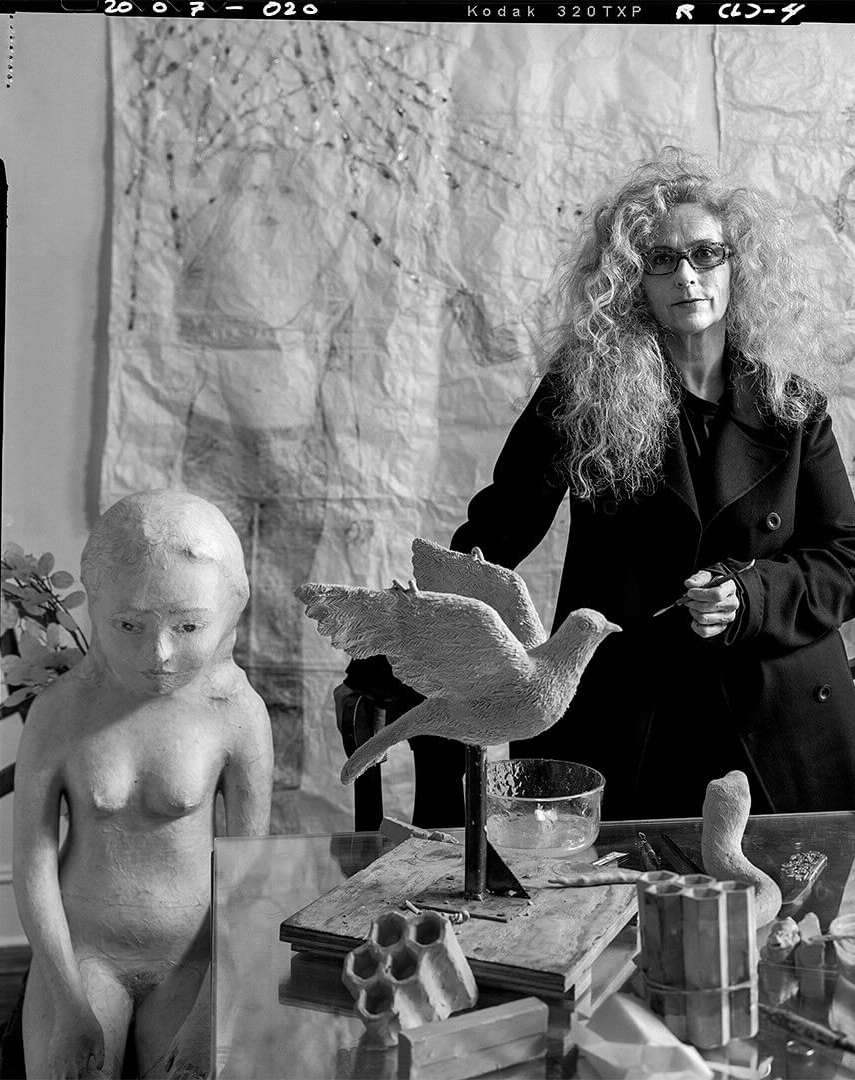 Black and white portrait of an adult woman with curly hair standing near a table with a bird sculpture and other art objects.