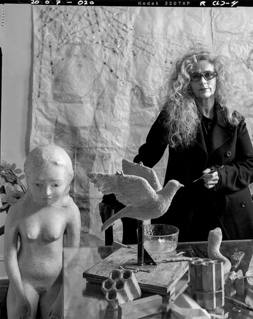 Black and white portrait of an adult woman with curly hair standing near a table with a bird sculpture and other art objects.