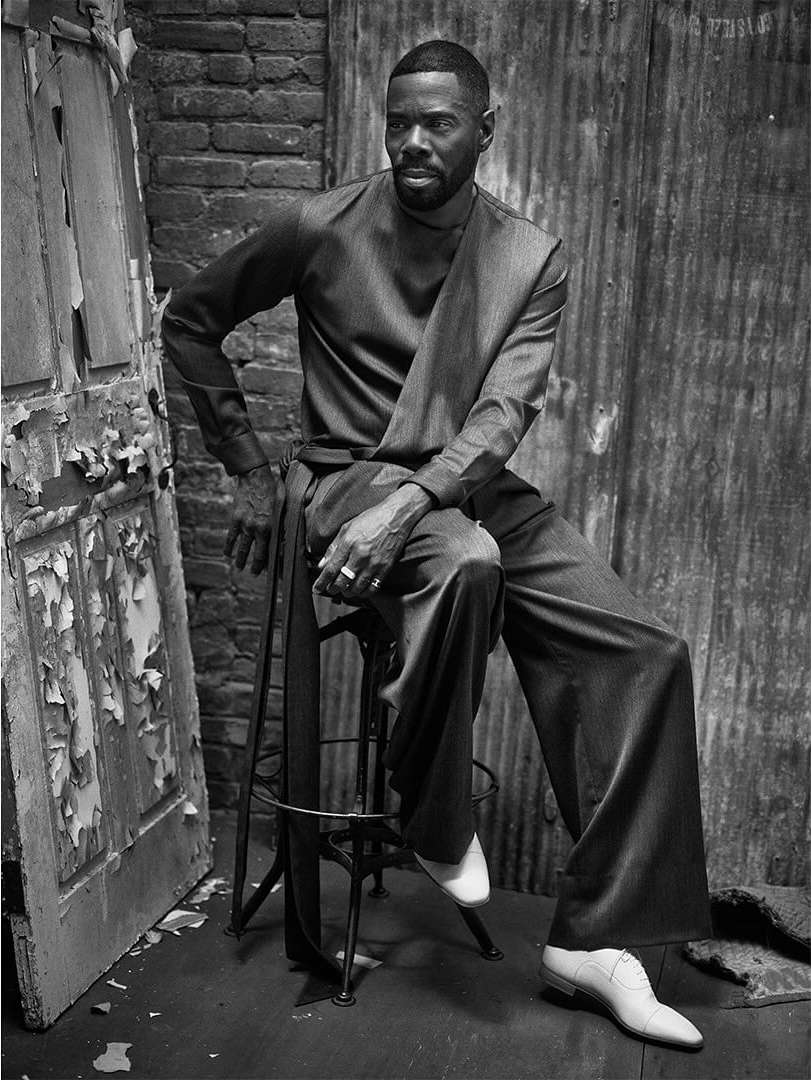 Monochrome portrait of a black man in an ash-grey suit seated on a stool in an urban location with a brick wall and peeling door.