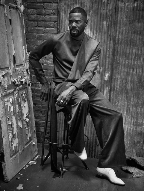 Monochrome portrait of a black man in an ash-grey suit seated on a stool in an urban location with a brick wall and peeling door.