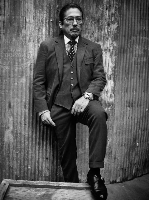 Portrait of a man in a suit leaning against a corrugated metal wall, wearing glasses and dress shoes, in black and white.