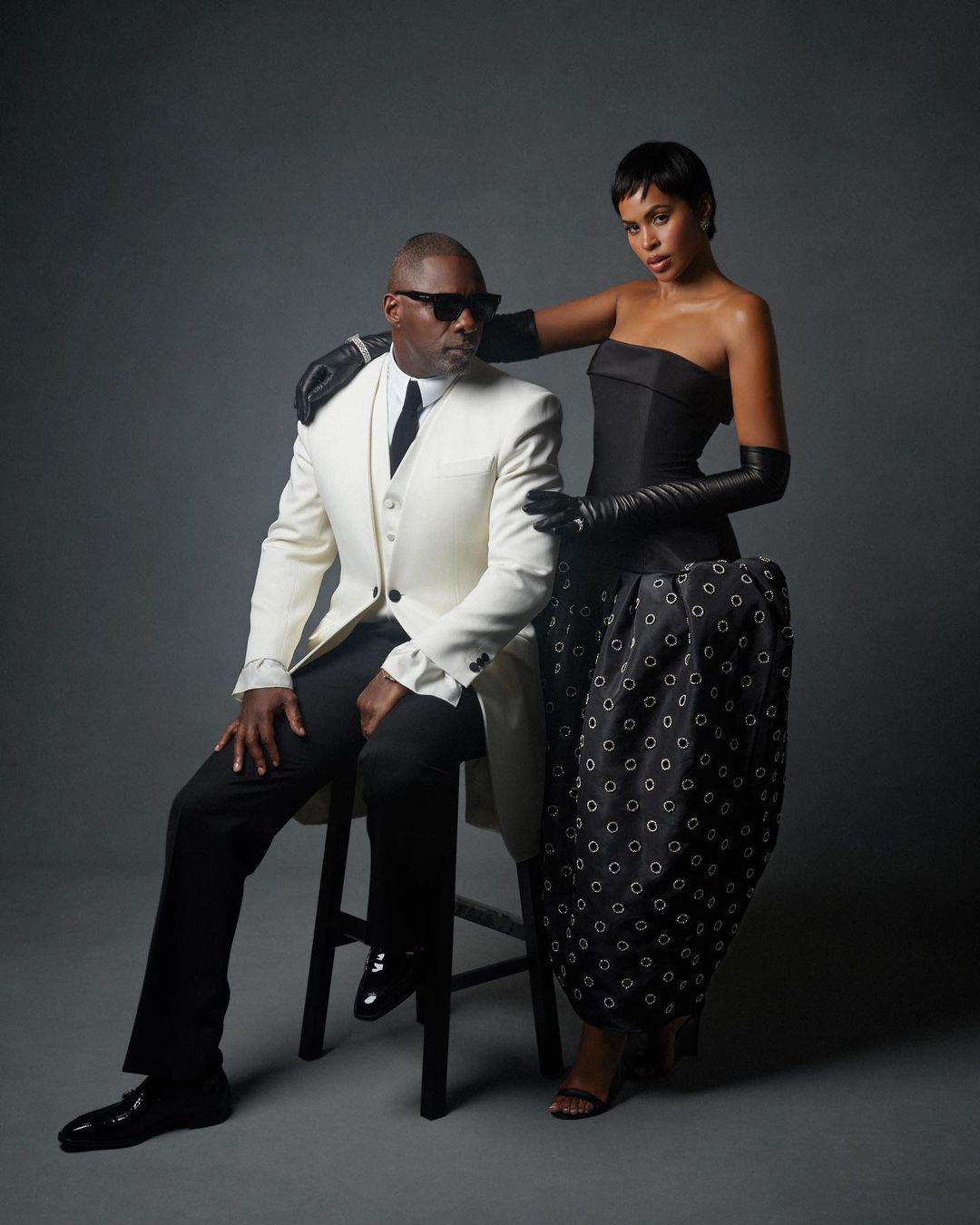 Elegant studio portrait of a Black adult male in a white suit seated on a stool, with a Black adult female in a black dress standing behind.