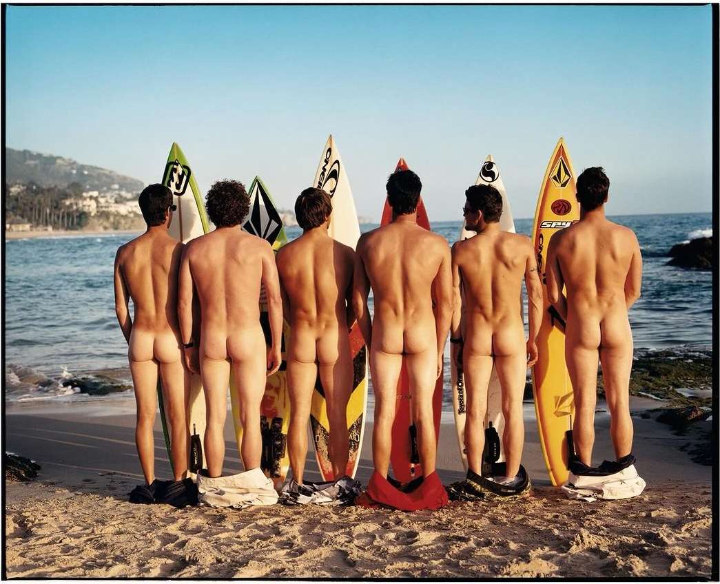 Six nude adult males on a beach stand in front of surfboards in sunny weather, facing away from the camera, with their clothing at their feet.