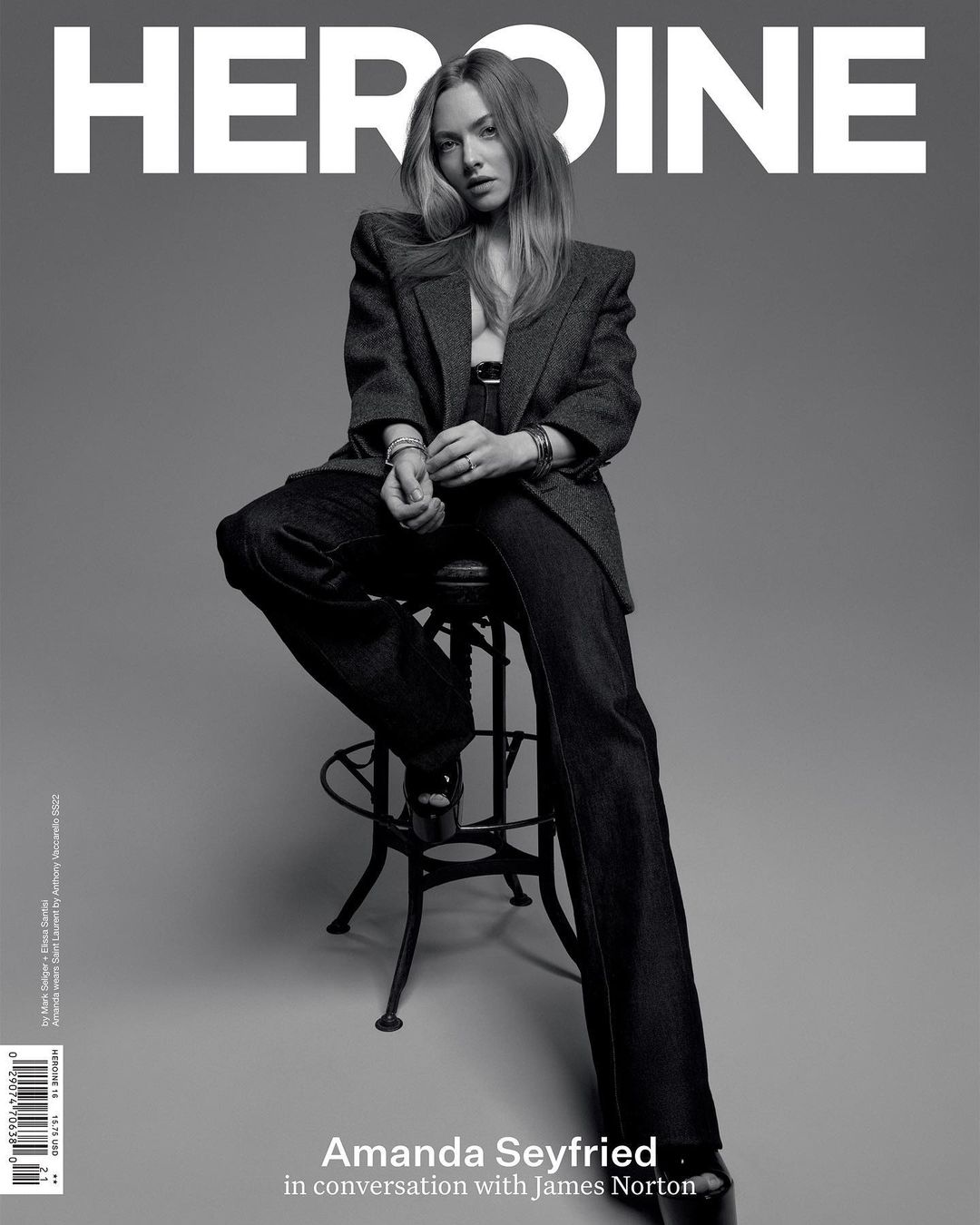 Amanda Seyfried sits on a stool in Heroine magazine cover, wearing a dark blazer, pants, and platform heels against grey backdrop.