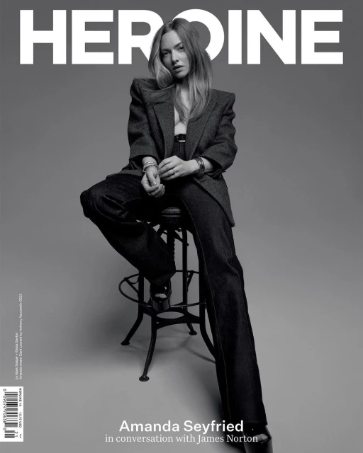 Amanda Seyfried sits on a stool in Heroine magazine cover, wearing a dark blazer, pants, and platform heels against grey backdrop.