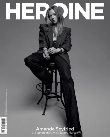 Amanda Seyfried sits on a stool in Heroine magazine cover, wearing a dark blazer, pants, and platform heels against grey backdrop.