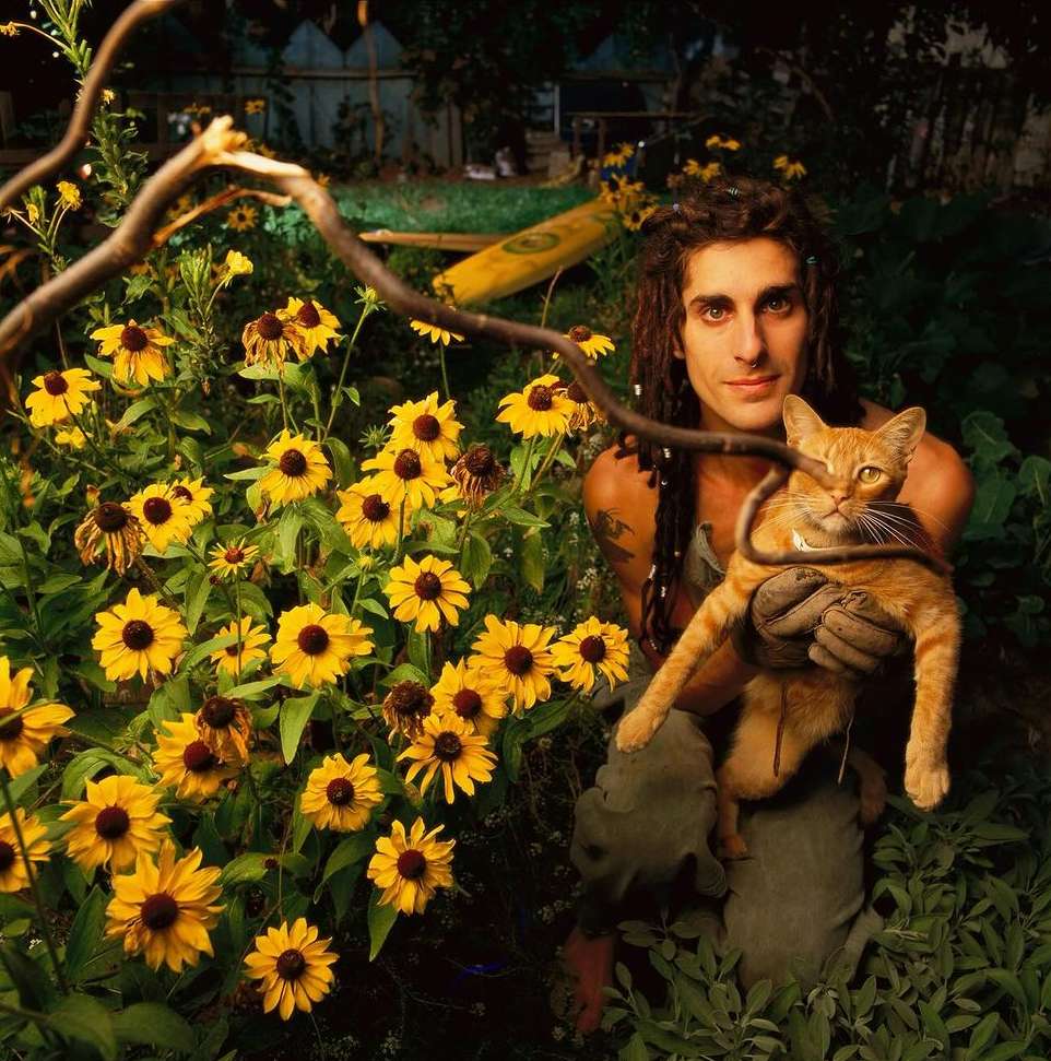 Man with dreadlocks cradles an orange tabby cat among yellow flowers in a lush green garden, featuring a rustic, natural setting.