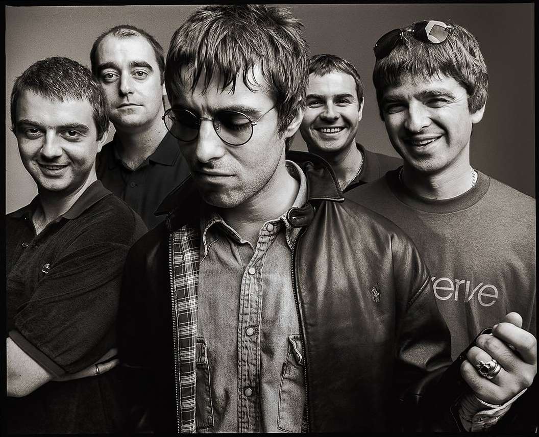 Black and white portrait of the band Oasis, featuring five adult males, leather jacket, button-down shirt, and casual attire in a studio setting.