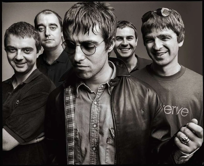 Black and white portrait of the band Oasis, featuring five adult males, leather jacket, button-down shirt, and casual attire in a studio setting.