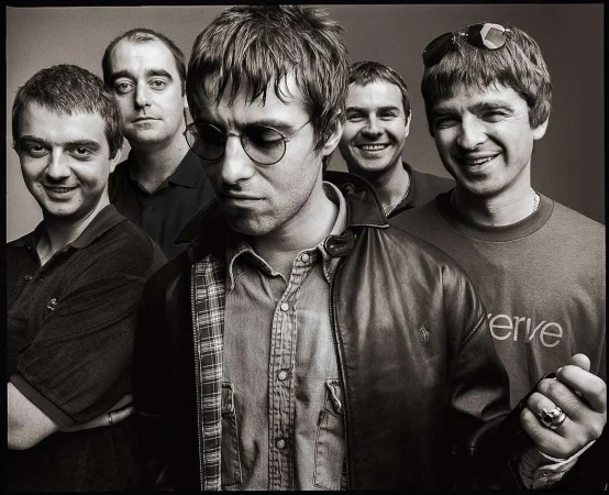 Black and white portrait of the band Oasis, featuring five adult males, leather jacket, button-down shirt, and casual attire in a studio setting.