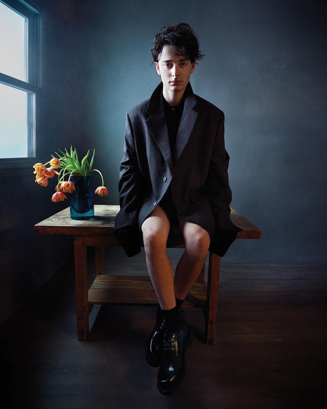 Portrait of a young adult with dark hair wearing an oversized blazer, seated on a rustic wooden table next to a vase of tulips.