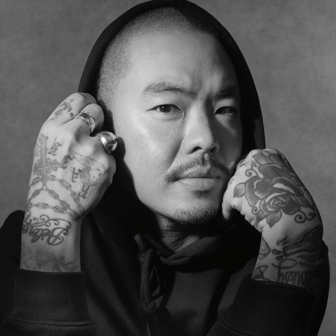 Striking black and white portrait of an adult man with tattoos, wearing a hooded sweatshirt, captured in a close-up studio setting.